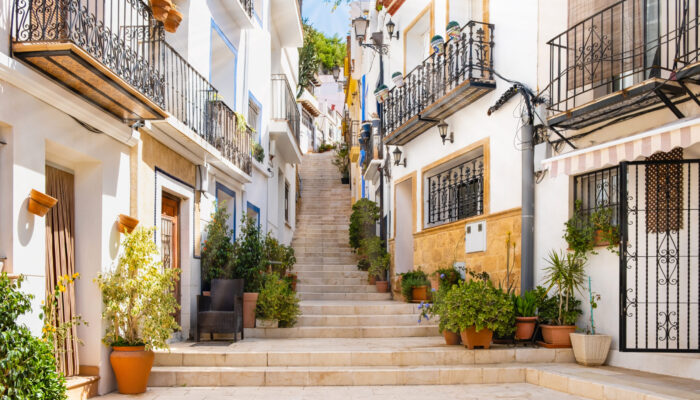 Narrow street with steps, white houses and potted plants in ancient neighborhood El Barrio or Casco Antiguo Santa Cruz in Alicante old town on hillside. Costa Blanca on Mediterranean sea coast, Spain